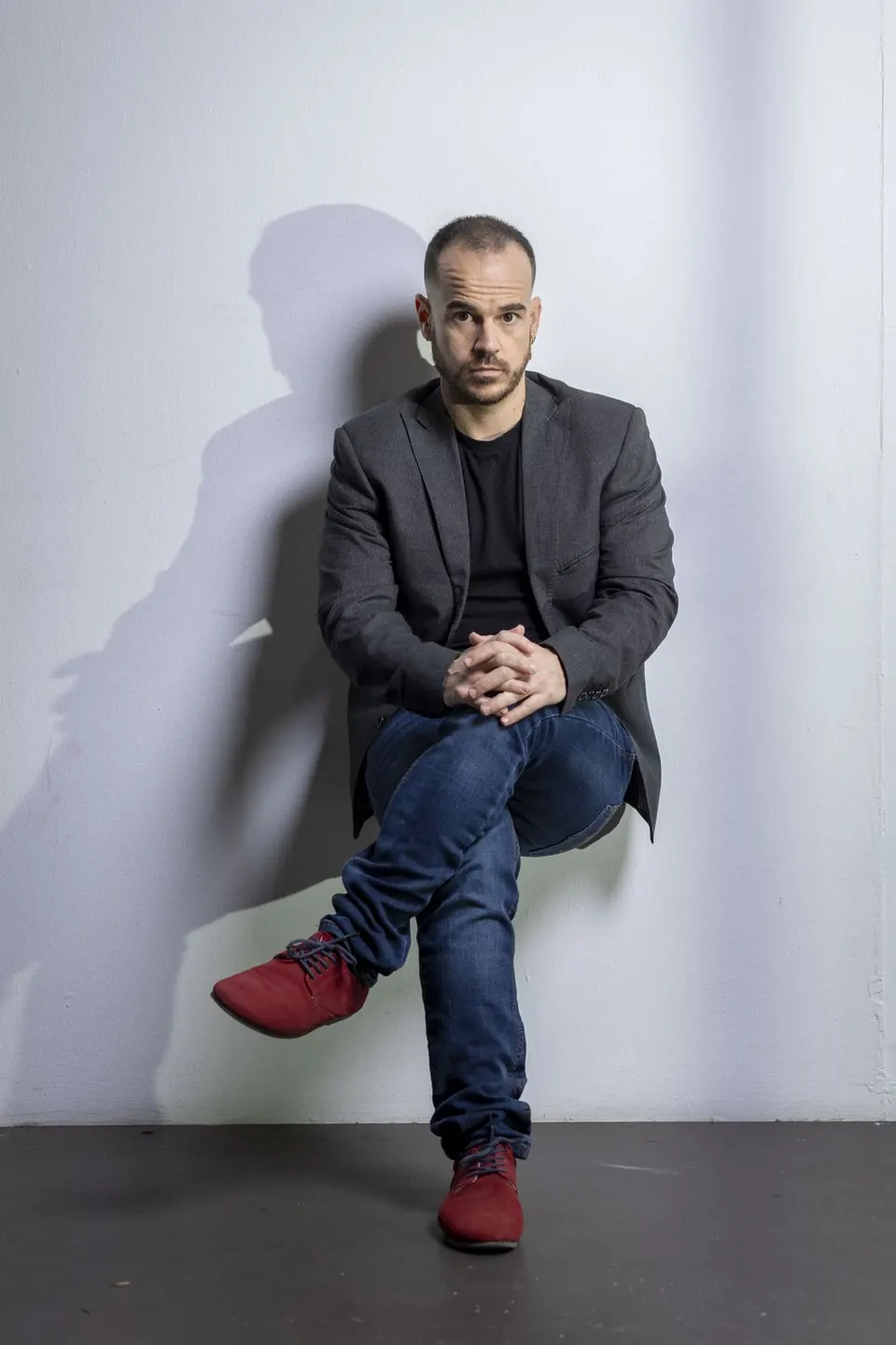 Charlie, con americana y zapatillas rojas, sentado en el aire contra una pared, sin silla