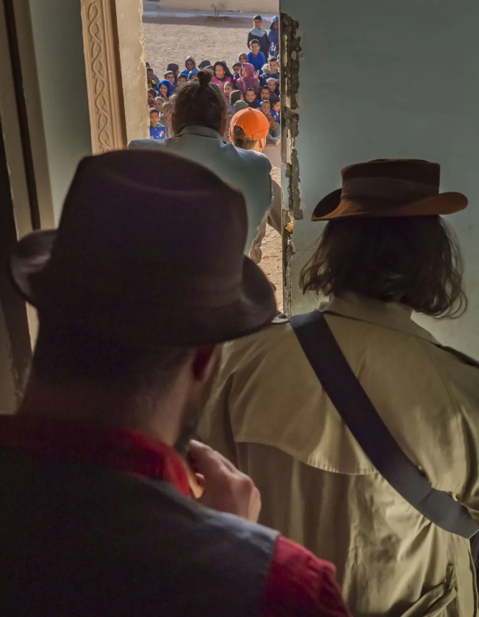Dos magos de espaldas, con sombrero, miran a los niños que esperan en fila al otro lado de una puerta